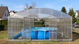 A clear arched pool enclosure with metal framing enclosing a blue above-ground swimming pool, set in a backyard with grass, shrubs, and houses in the background.