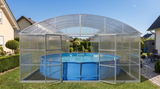 A large arched aluminum pool enclosure with clear panels enclosing an above-ground blue pool, shown in a backyard setting with grass, shrubs, and neighboring houses visible.