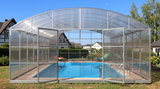 A large arched pool enclosure with a transparent screen wall and frame, enclosing a blue swimming pool in a backyard setting with grass, trees, and houses visible in the background.