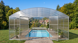 A large arched transparent pool enclosure with glass panels and open side doors, enclosing a rectangular swimming pool on a wooden deck, set in a grassy backyard with trees and a distant village under a blue sky.